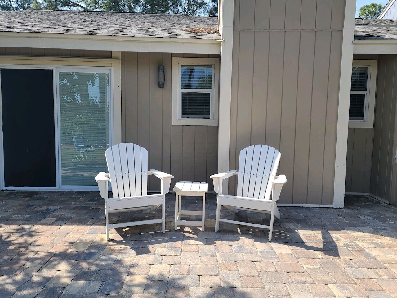 A patio area with two white Adirondack chairs and a small table, in front of a building with beige siding and a sliding glass door.