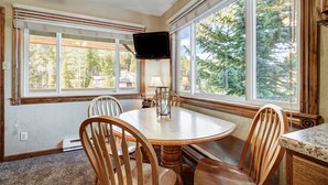 A dining area with a round wooden table, four chairs, a TV mounted on the wall, and large windows offering a view of trees. - Cozy, Corner Top Floor Unit in Perfect Downtown Location | PM8B (Breckenridge)