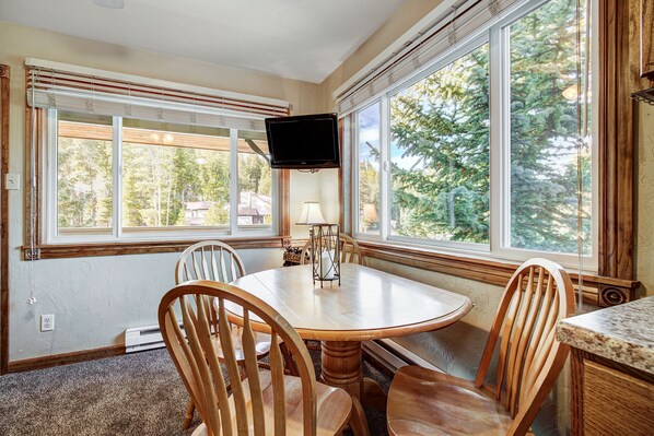 A dining area with a round wooden table, four chairs, a TV mounted on the wall, and large windows offering a view of trees.