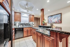 A kitchen with wooden cabinets, granite countertops, a dishwasher, and a ceiling fan. - Kihei Beach #409 – Immersive Views and Tranquil Escape (Kihei, Maui)
