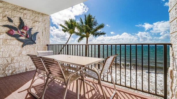 A balcony with a table and chairs, a decorative wall, and a view of the sea and palm trees. - Kihei Beach #409 – Immersive Views and Tranquil Escape (Kihei, Maui)