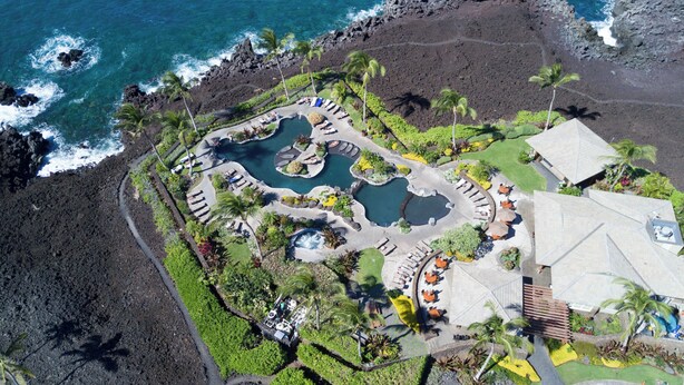 Luftaufnahme eines Resorts mit mehreren Schwimmbecken, einem großen Gebäude und einer umliegenden, bepflanzten Anlage mit Palmen.