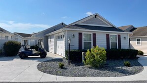 A single-story house with a garage, a small garden with gravel and plants, and a golf cart parked in front. - Walking distance to Lake Sumter Landing! Small Dog Friendly Villa w/Golf Cart (The Villages)