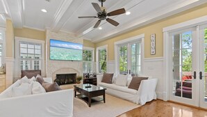 A spacious living room with a fireplace, a ceiling fan, and large windows.