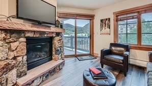 Un salon doté d’une cheminée en pierre, d’un téléviseur à écran plat, d’un fauteuil en cuir et offrant une vue sur un balcon avec une balustrade en bois.