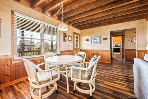 A wooden interior with a round table and chairs, a kitchen in the background, and a view through large windows.