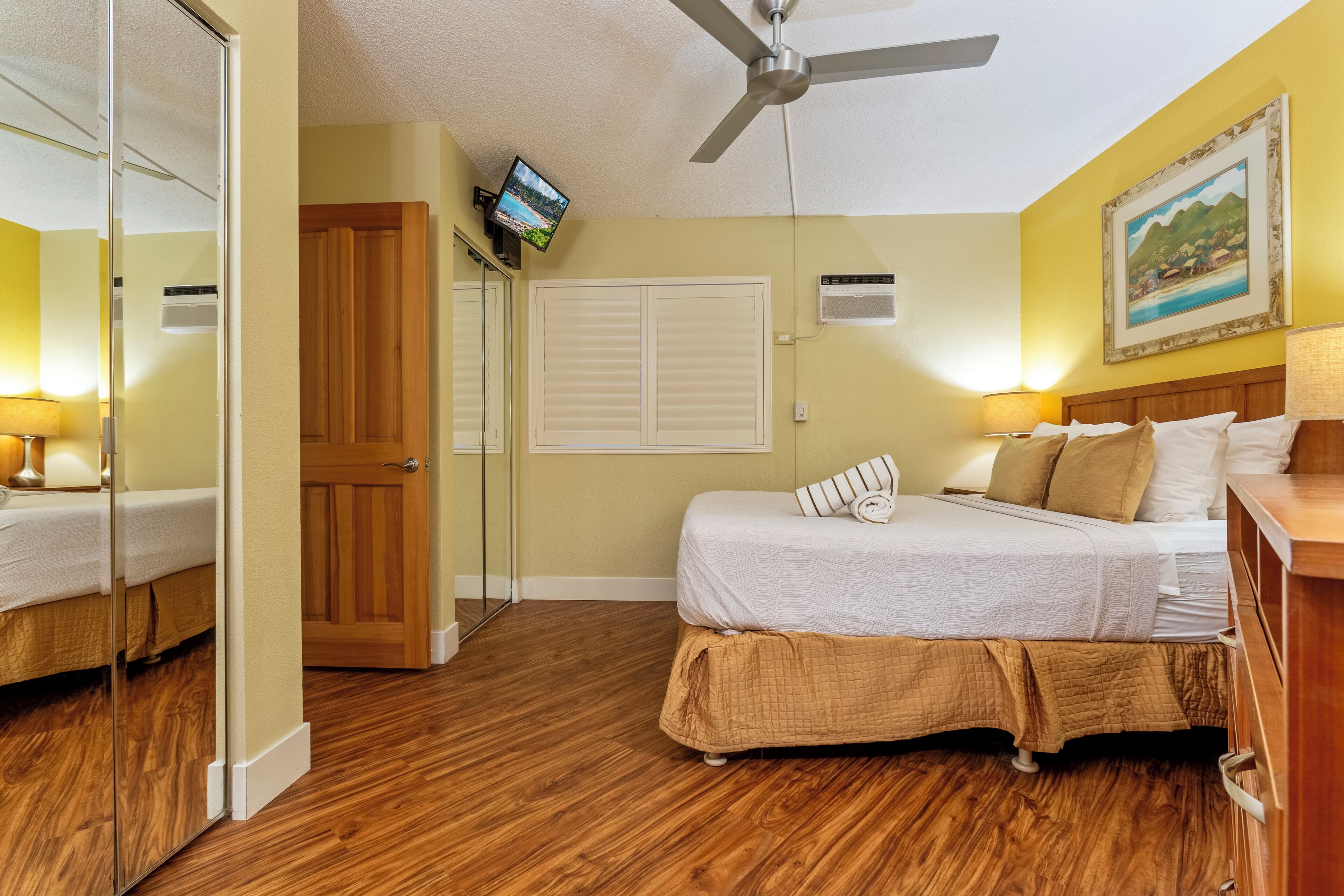 A bedroom with a bed, wooden floor, ceiling fan, TV, and a painting on the wall.
