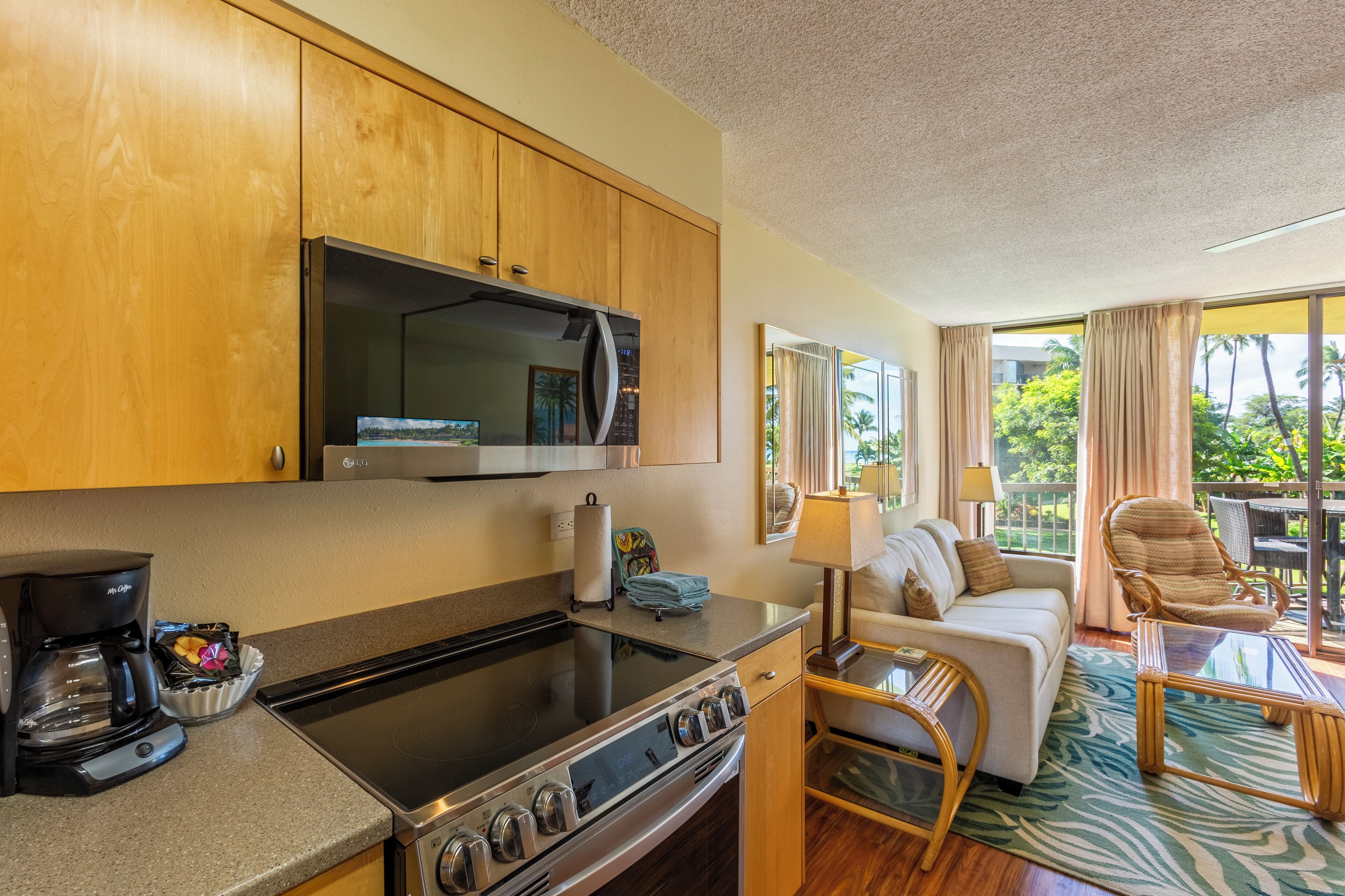 A compact kitchen with a stove, oven, and coffee maker, a sofa, and a chair in a room with large windows and a view of greenery.