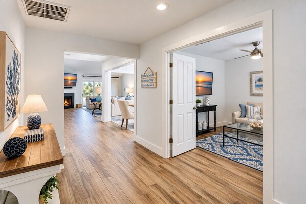 Een ruime woonkamer met houten vloer, een zithoek met een bank en fauteuils, een salontafel en een aan de muur bevestigde televisie.