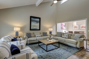 A spacious living room with a large sectional sofa, a coffee table, and two side tables with lamps. There is a large window providing natural light and artwork on the walls.