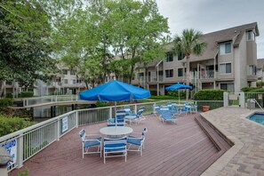A deck with outdoor seating and umbrellas, a pool, and a building with balconies.