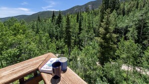 Una mesa de picnic con un libro y dos tazas, con vistas a un bosque frondoso y montañas.