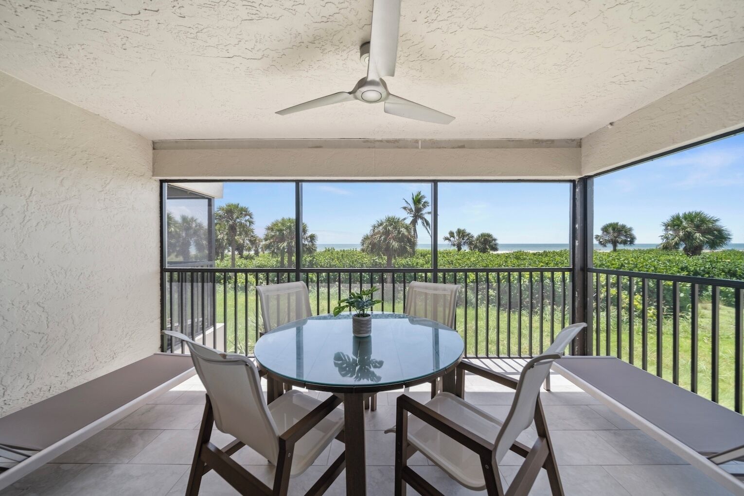 A balcony with a glass table and chairs, a ceiling fan, and a view of the ocean and palm trees.