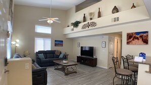A spacious living area with a ceiling fan, a flat-screen TV, a dark-colored sofa, a coffee table, and a dining area with metal chairs. - Home Field Advantage by Mesquite Vacation Rentals with Community Pool and $510 in free daily activities (Mesquite)