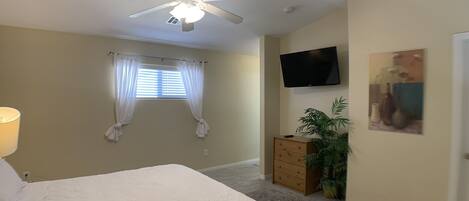 Una camera da letto con un letto, una ventola a soffitto, una televisione, un comò e una finestra con tende.