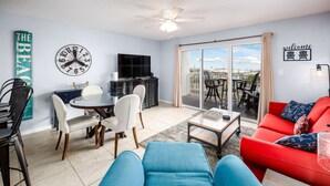 A modern living room with a blue sofa, a red sofa, a dining area with a table and chairs, a TV, and a balcony with a view.