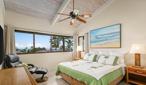 Ein Schlafzimmer mit einem Bett, einem Schreibtisch, einem Sessel, einem Deckenventilator und einem großen Fenster mit Blick auf den Strand.