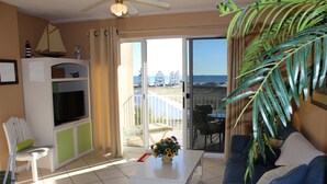 A living room with a television, a sofa, a coffee table with flowers, and a view of the beach through a sliding glass door.