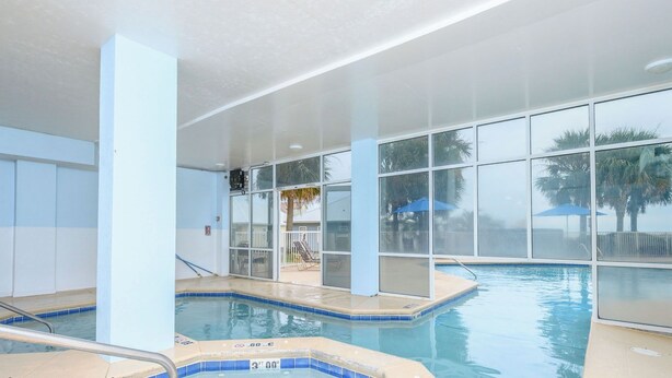 Une piscine intérieure avec des parois en verre transparent, un jacuzzi et une vue sur les palmiers à l’extérieur.