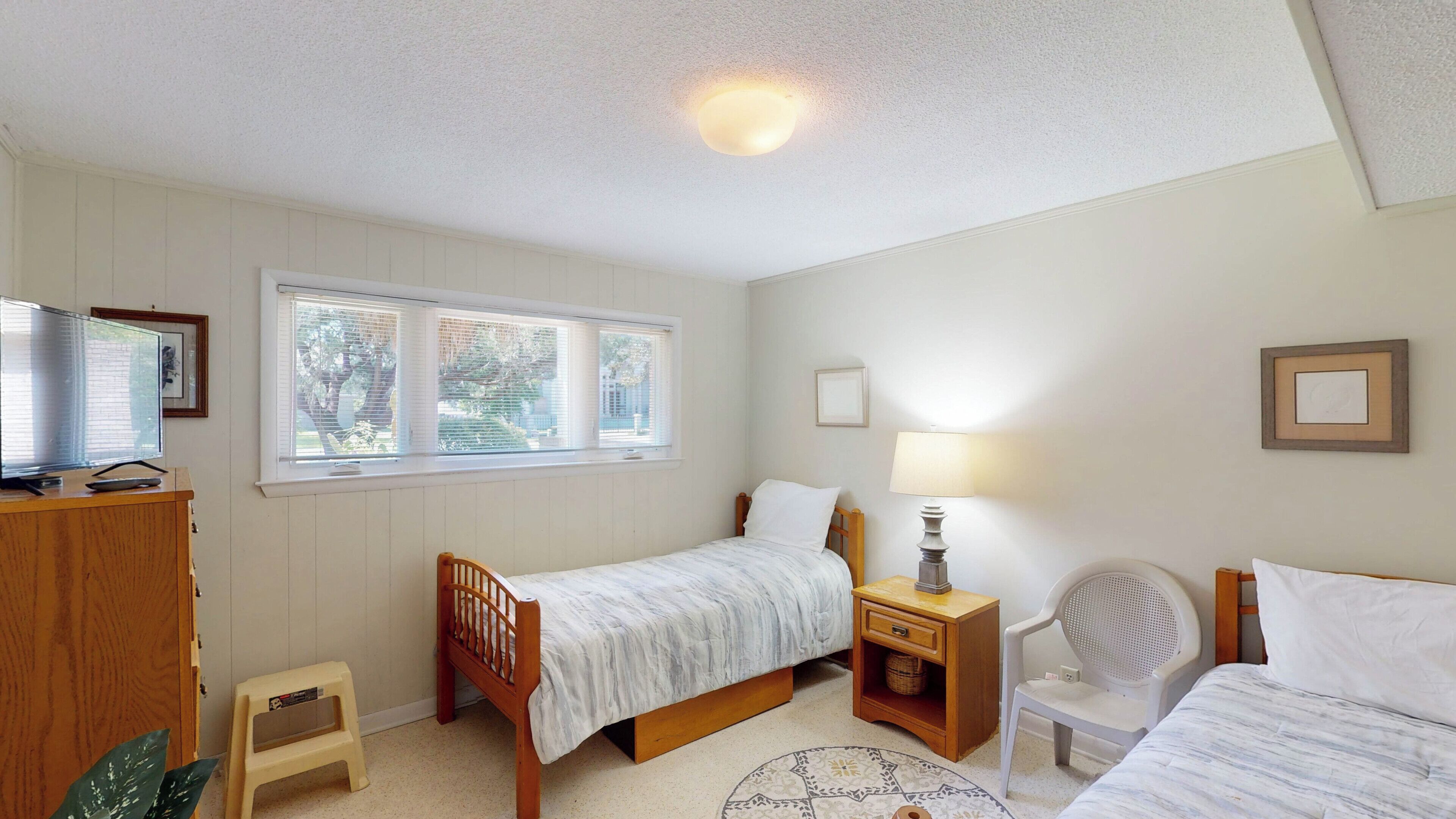 A bedroom with two single beds, a wooden nightstand, a chair, a small stool, and a television on a wooden cabinet.