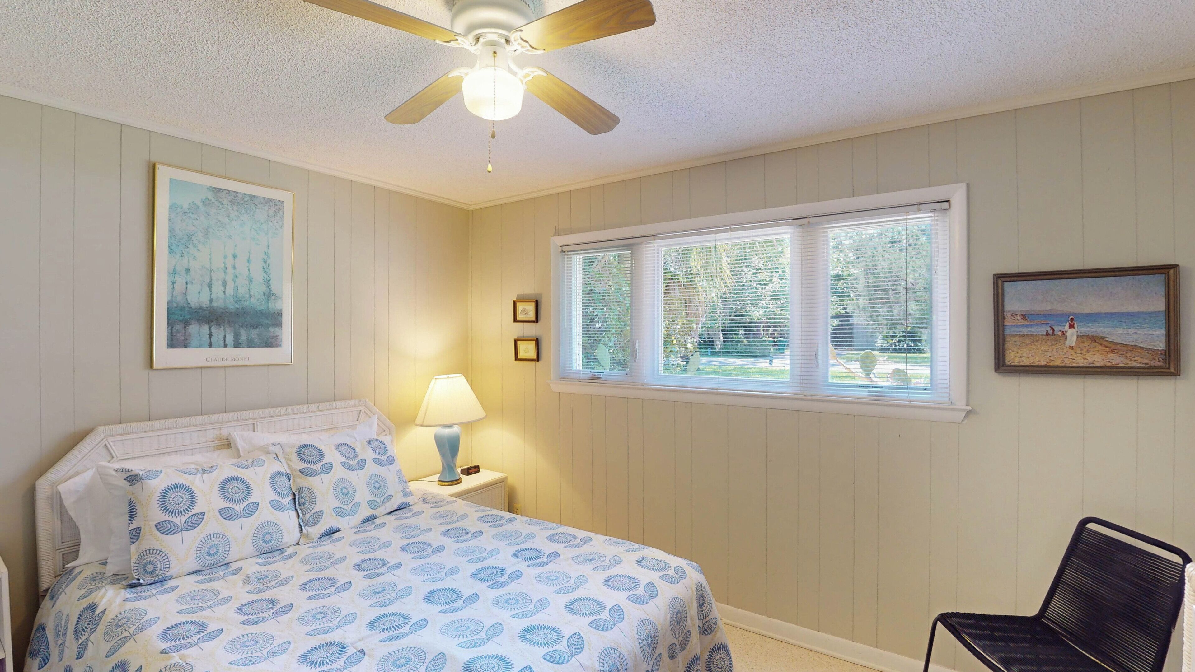 A bedroom with a bed, a nightstand, a lamp, a ceiling fan, a window with blinds, and two framed pictures on the wall.
