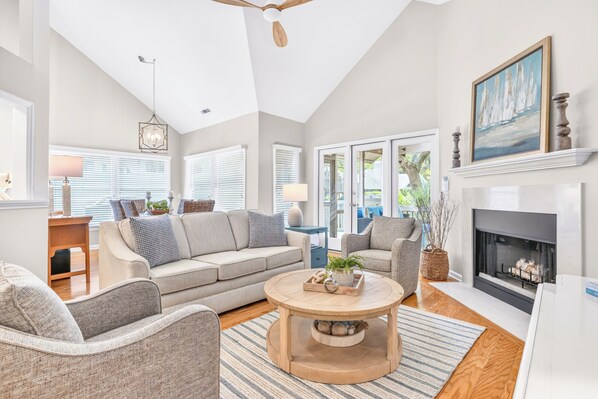 A spacious living room with a fireplace, a ceiling fan, and a large window with blinds.