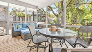 Een overdekt terras met een ronde glazen tafel, rieten meubels en uitzicht op omliggende huizen.