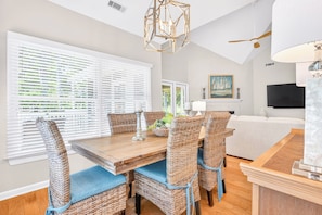 A dining area with wicker chairs and a wooden table, a white sofa, a ceiling fan, and a painting on the wall.