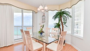 A dining area with a glass table, white chairs, a potted plant, and a view of the ocean through large windows. - Beachfront Condo. Stunning Views from Balcony. (Destin)