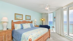 A bedroom with a large bed, wooden headboard, two bedside tables with lamps, a small table with a lamp, and a view of the ocean through large windows. - Beachfront Condo. Stunning Views from Balcony. (Destin)