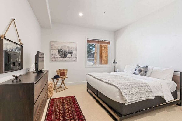 Une chambre à coucher avec un lit, une commode, un téléviseur, une fenêtre et un tapis.