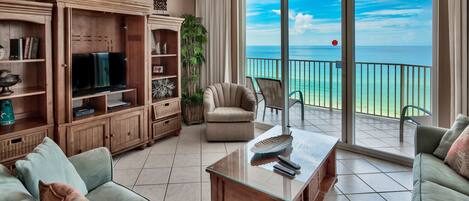 A living room with a TV, bookshelves, a sofa, and a balcony with a view of the ocean.