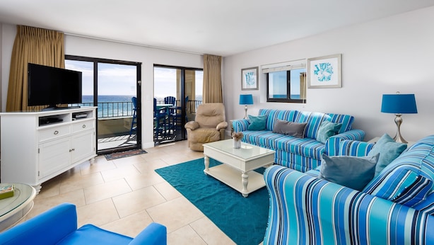 A living room with a sofa, armchair, and television, a balcony with chairs, and a view of the ocean. - Surf Dweller 312🌊Gulf Front🏖️Free Beach Setup (Fort Walton Beach)