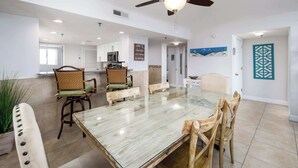 A spacious dining area with a glass-top table and wooden chairs, a ceiling fan, and a kitchen in the background. - Surf Dweller 312🌊Gulf Front🏖️Free Beach Setup (Fort Walton Beach)