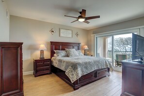 A bedroom with a large bed, two bedside tables, a ceiling fan, and a large window with a view.
