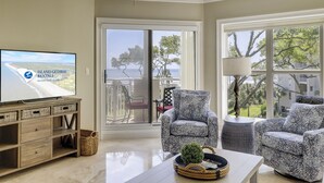 A spacious living room with a television displaying 'Island Getaway Rentals' and a scenic view of the ocean through large windows. - 1 Bedroom/2 Bathroom 3rd Floor Oceanfront End Unit Condo with Amazing Views!! (Hilton Head Island)