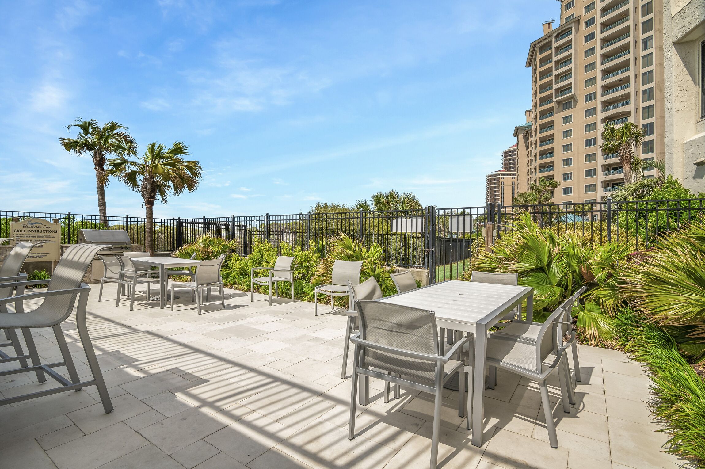 An outdoor patio area with tables and chairs, surrounded by palm trees and greenery, with a high-rise building in the background.