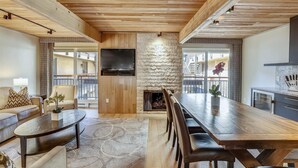 A spacious living area with a long wooden dining table, a fireplace, and a TV mounted on a stone wall. - Elegant Downtown Chateau | Walk to Shop, Ski & Dine (Aspen)
