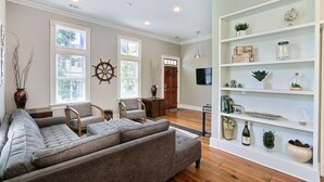 A living room with a grey sectional sofa, two armchairs, a wooden coffee table, a ship's wheel decoration, a bookshelf with various items, and a door leading to another room. - Coastal Townhome Near Park with Parking by Lucky Savannah (Savannah)