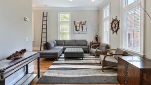 A living room with a sectional sofa, ottoman, and wooden coffee table. There are two windows with blinds, a ladder, and a wall-mounted ship's wheel. - Coastal Townhome Near Park with Parking by Lucky Savannah (Savannah)