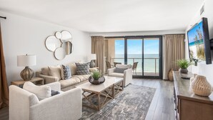 A spacious living room with a large window offering a view of the sea, a sofa set, a coffee table, and a TV mounted on the wall. - Golf Cart ~ Gulf Views ~ Private Balcony (Miramar Beach)