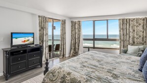 A bedroom with a large bed, a TV on a black cabinet, and a view of the ocean through large windows. - Golf Cart ~ Gulf Views ~ Private Balcony (Miramar Beach)