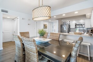 A modern kitchen with stainless steel appliances, a dining area with wicker chairs, and a large pendant light. - Golf Cart ~ Gulf Views ~ Private Balcony (Miramar Beach)