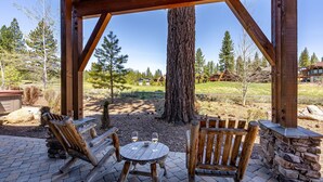 Une terrasse extérieure en bois, avec une table et des chaises en pierre, donnant sur une zone verdoyante avec des arbres et un bâtiment situé au loin.
