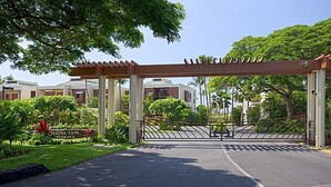 Une entrée fermée par un portail, agrémentée d’une pergola, une enseigne indiquant « Mauna Lani Terrace », et une vue sur des immeubles résidentiels en arrière-plan.