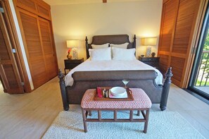 Quarto com uma cama grande, duas mesas de cabeceira com luminárias, um banco de madeira e vista pela janela.