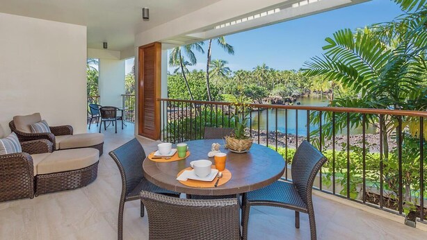 A balcony with wicker furniture, a dining table set with cups and a bowl, and a view of a tropical landscape with palm trees and a body of water.