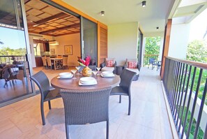 Uma varanda com móveis de vime, uma mesa de jantar posta com pratos e copos, e vista para o exterior.