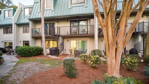 A multi-story residential building with a green roof, outdoor seating area, and a tree in the foreground. - Surf Court 62 - Forest Beach 1st Floor Flat (Hilton Head Island)