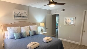 A bedroom with a bed, pillows, a nightstand, a lamp, a ceiling fan, and artwork on the walls. - Surf Court 62 - Forest Beach 1st Floor Flat (Hilton Head Island)